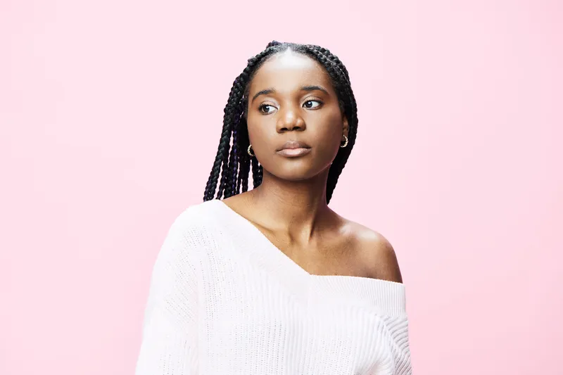 Young Black woman with braids in an off shoulder white sweater against a pink background, conveying a feeling of calm and confidence