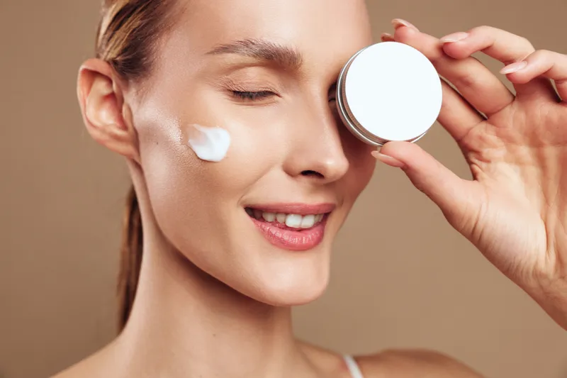Young woman applies cream to her cheek in a close up shot against a beige background, conveying skincare routine, self care and a calm, glowing mood.