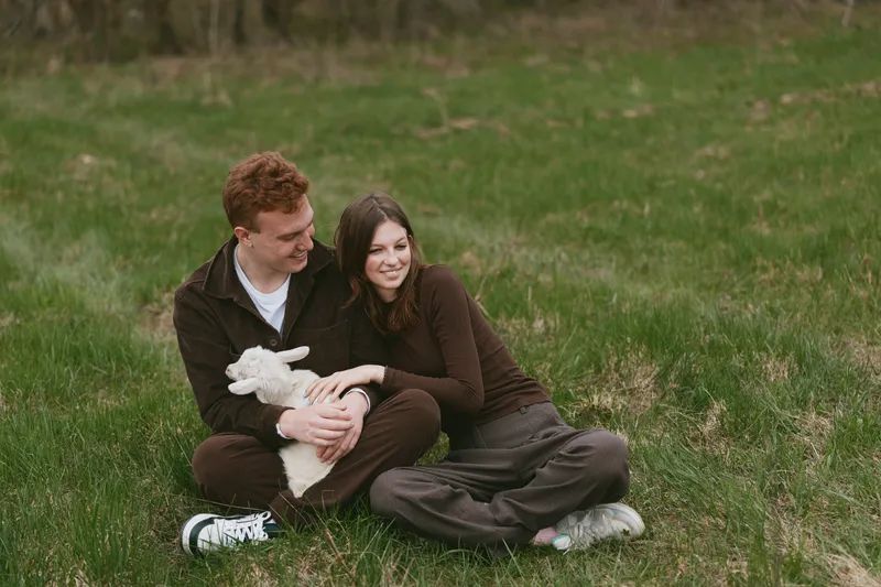 Couple outdoors sharing affection in a park on grass with a lamb, love and togetherness, warm smiles, serene nature, candid lifestyle portrait, daytime happiness and cozy bond