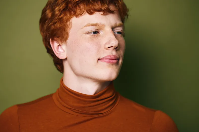 Portrait of a young man with red hair wearing a brown turtleneck sweater looking sideways against a green background. Closeup of face and thoughtful expression in studio lighting.