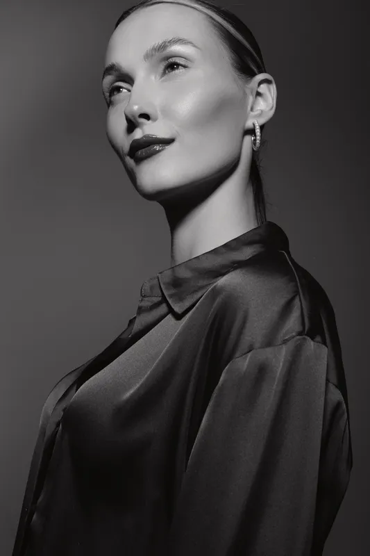 Portrait of a pale white woman with slicked hair wearing silk blouse and earrings looking away thoughtfully in black and white studio. Concept of elegance, fashion, and beauty.
