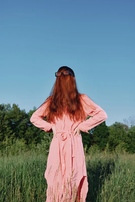 Young woman in a pink dress standing in a green field, showcasing her long red hair against a clear blue sky, embodying a relaxed and carefree attitude