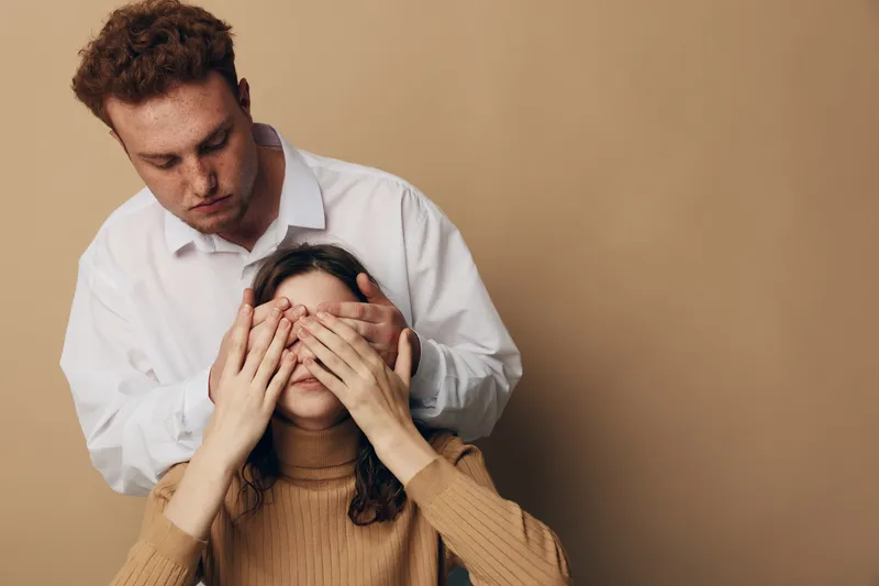 Young man covers eyes of woman from behind in beige studio. Couple plays surprise game with gentle touch and warm casual clothing. Concept of love, trust, and relationship.