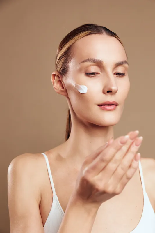 Young woman applying white face cream on cheek with closed eyes on beige background. Concept of skincare, beauty, wellness, and self-care with soft natural lighting.