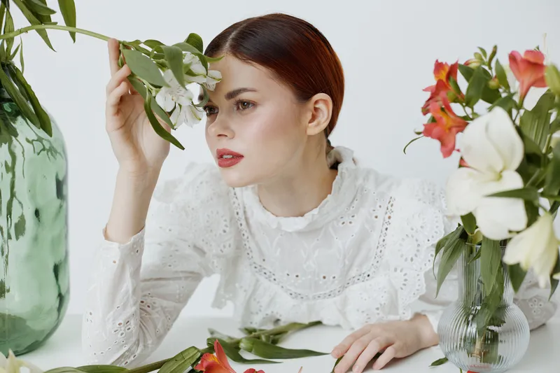 A young woman with red hair in a white lace blouse surrounded by vibrant flowers, conveying freshness and tranquility in a minimalist setting