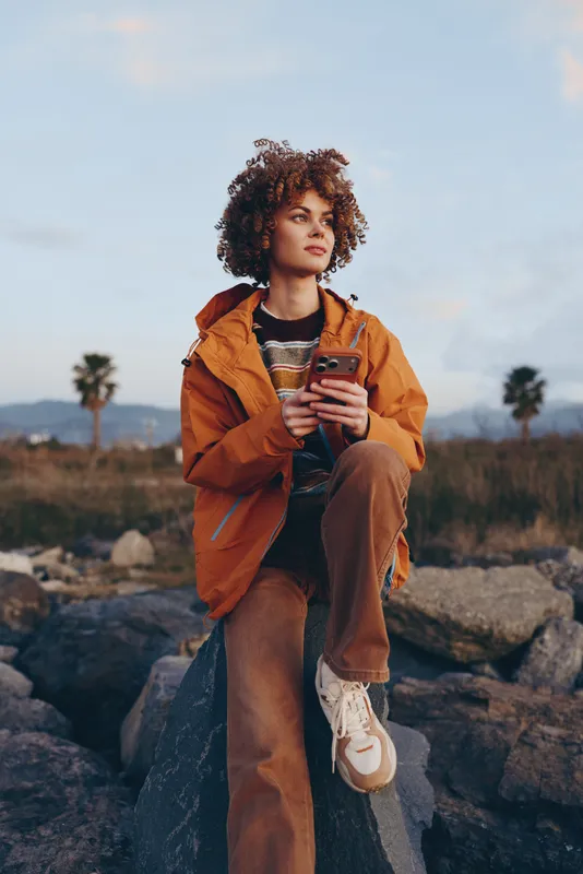 Woman lifestyle portrait outdoors, curly hair orange jacket, holding a smartphone, standing on rocks, palm trees and mountains in background, travel mood, soft cinematic light