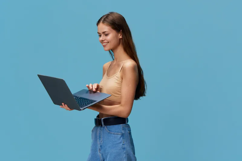 smiling woman using laptop with casual outfit and blue background, focused on screen and typing, young adult female working remotely or studying