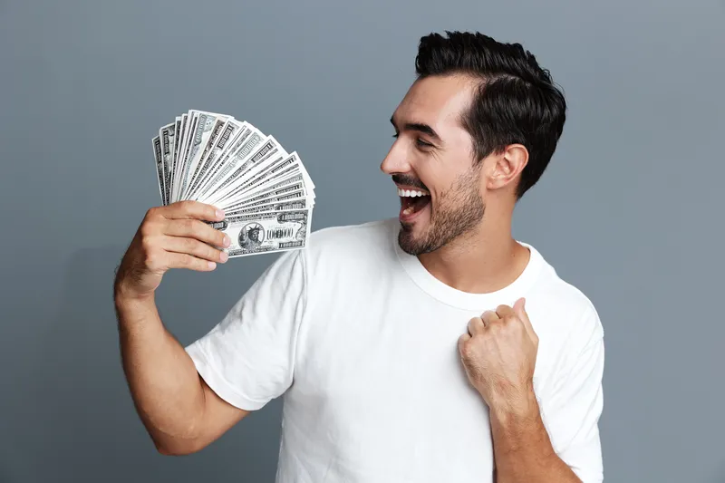 Happy smiling man in white t-shirt looks excited while holding a fan of cash in hand, isolated on solid grey background. Finance and lifestyle concept.