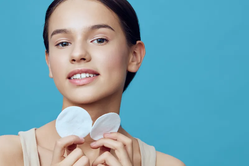 Young woman with soft skin holding cotton pads, smiling against a blue background, representing beauty, skincare, and self care routines for healthy skin
