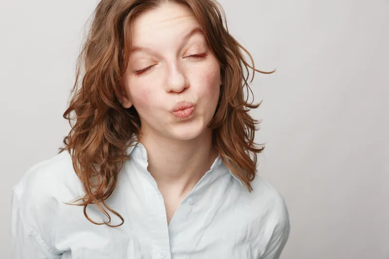 Young woman with curly hair wearing white shirt makes a playful kiss face with closed eyes. Studio portrait on light background. Concept of fun, youth, and casual lifestyle.