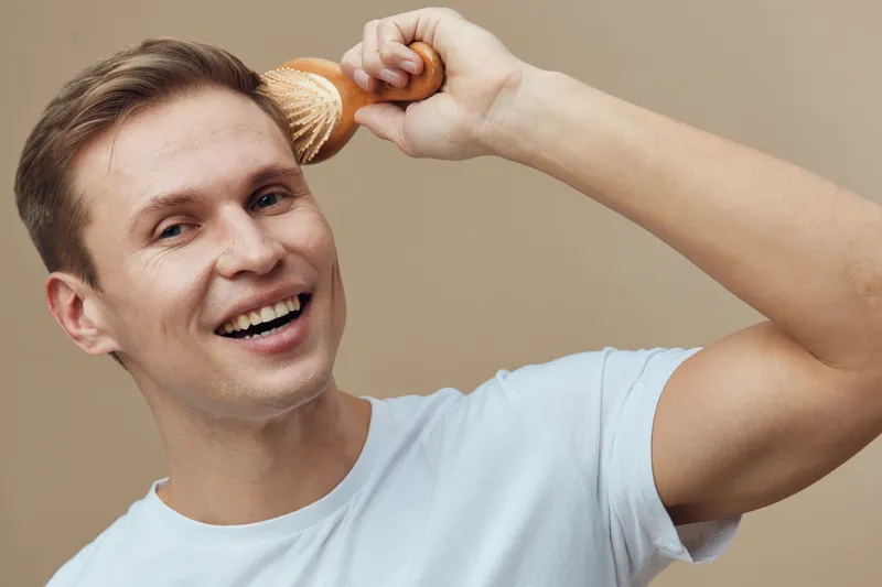 Smiling young man with light hair holding makeup brush near head, looking cheerful and confident, wearing casual white t-shirt, indoor setting with plain beige background, lifestyle concept, expressiv
