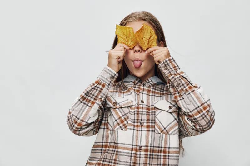 Girl in plaid shirt holding two autumn leaves over eyes and sticking out tongue with playful expression on light background. Child playing with nature and having fun indoors.