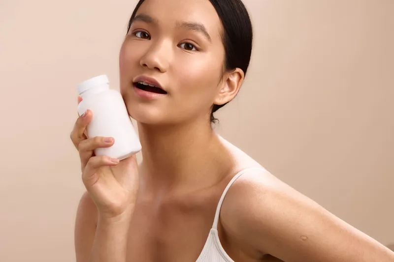 Asian woman holds a cosmetic bottle in a soft studio setting, smiling with confidence, creating a calm, approachable beauty moment that highlights a simple skincare routine.