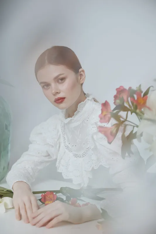 Elegant young woman in a white lace blouse surrounded by colorful flowers, showcasing a serene expression and soft pastel tones, ideal for seasonal fashion imagery