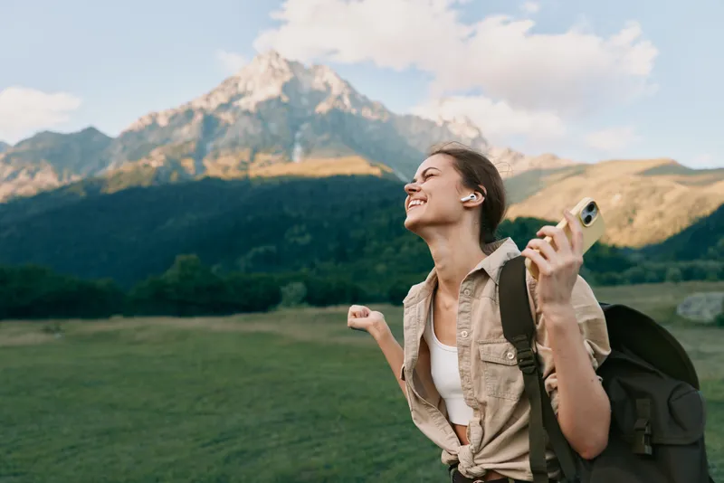Happy woman with backpack enjoying nature and listening to music with wireless earbuds in a mountain landscape. Lifestyle concept with freedom and relaxation outdoors.