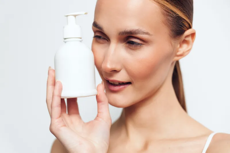 Beautiful woman holding a white pump bottle near her face, skincare routine, focused smile, soft studio light, clean background, moisturizing hydration, cosmetic care, hygiene