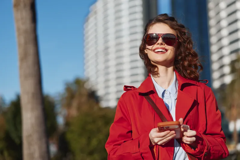 Joyful woman outdoors wearing a red coat and sunglasses, smiling while using a phone, sunlit urban scene, natural colors, relaxed vibe