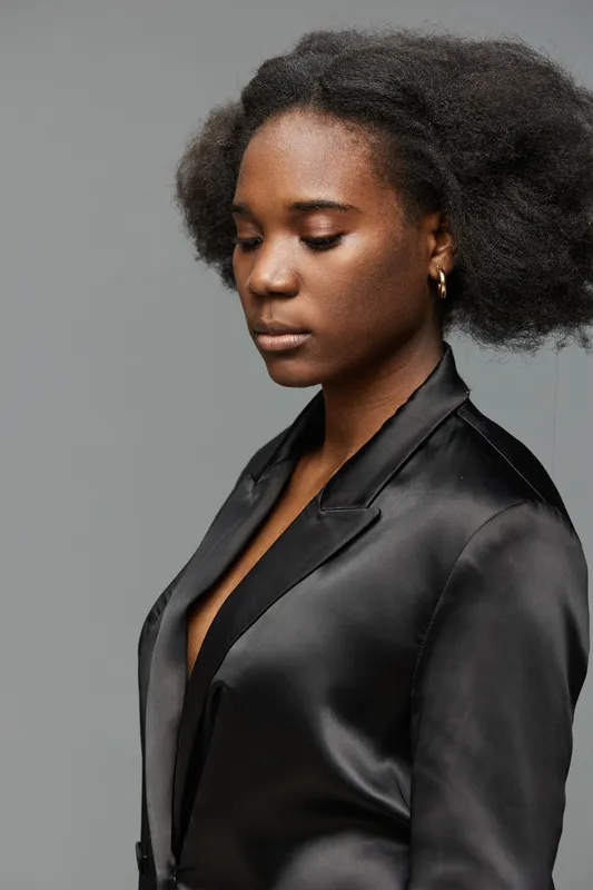 Elegant young woman with natural hair in a sleek black blazer, showcasing confidence and poise against a minimal gray background, perfect for fashion and beauty themes