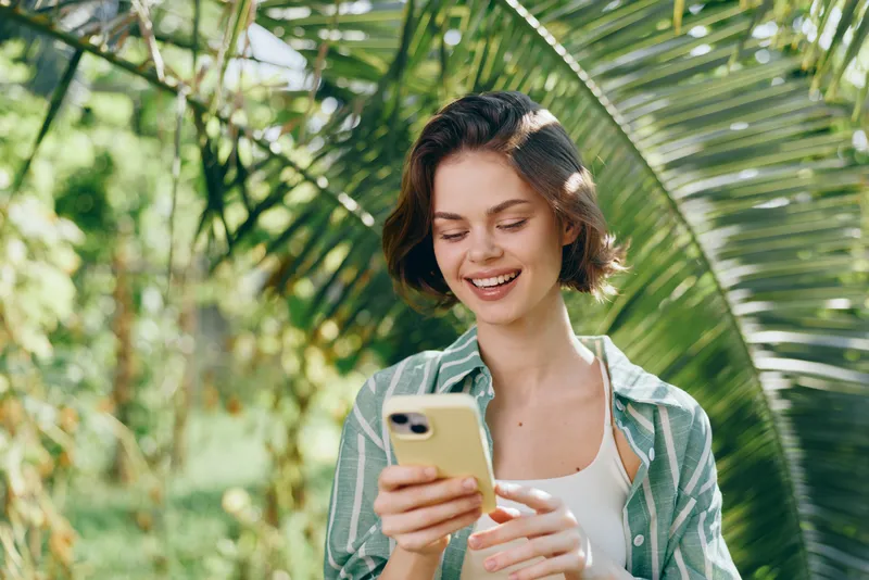 Young woman outdoors, smiling while using a smartphone amid tropical greenery, sunny and relaxed mood.