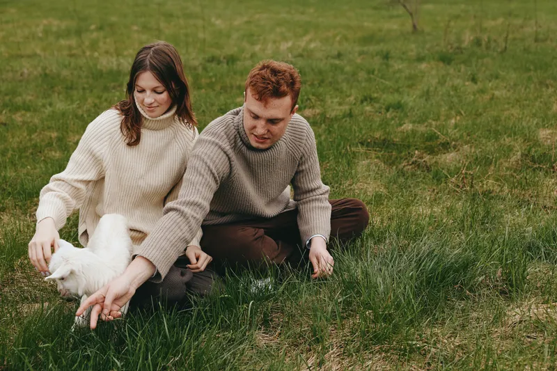 A couple in cozy sweaters interacts with a fluffy goat kid outdoors, sharing a gentle moment on a grassy field, capturing a warm lifestyle scene with countryside charm