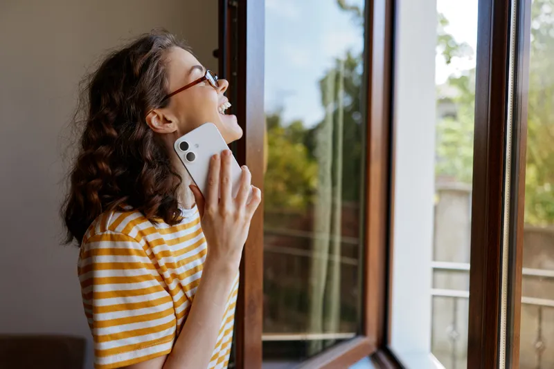 A cheerful woman in a yellow striped shirt talks on a smartphone by an open window, enjoying daylight and sunshine. A warm home moment capturing everyday connection and happiness.
