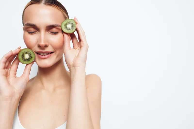 Young woman with closed eyes holding two halves of fresh kiwi fruit near her face. Clean white background, natural beauty concept, healthy lifestyle, and fresh organic skincare.