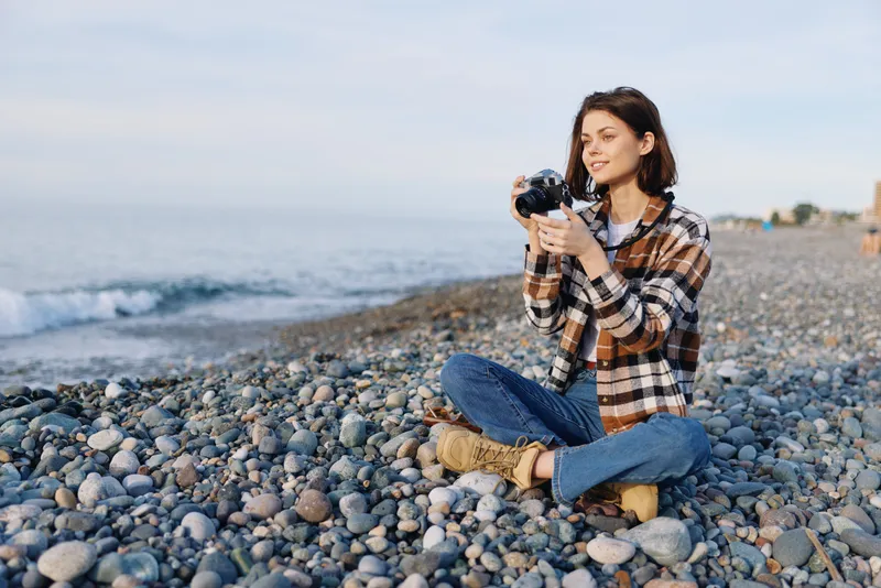 Woman photographer sits on a pebble beach, holding a camera and capturing seaside scenery in a casual outdoor lifestyle moment, sunlit and relaxed.
