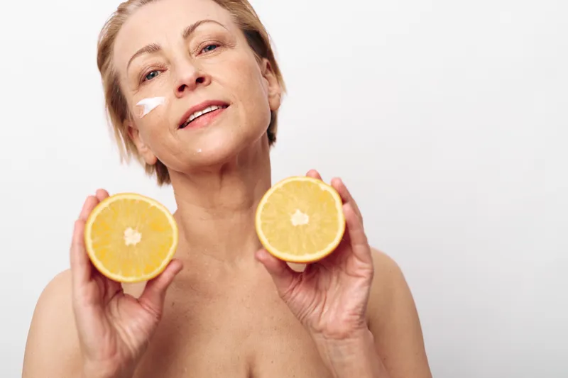 Mature woman with natural skin holds two orange halves close to face with cream on cheek. Concept of healthy skincare, vitamin C, natural beauty, and self-care routine in bright studio.