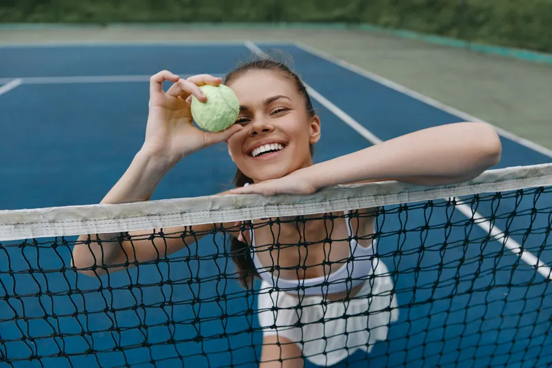 Joyful female tennis player holding a tennis ball, smiling with enthusiasm on a blue court background, emphasizing sport, happiness, and fitness