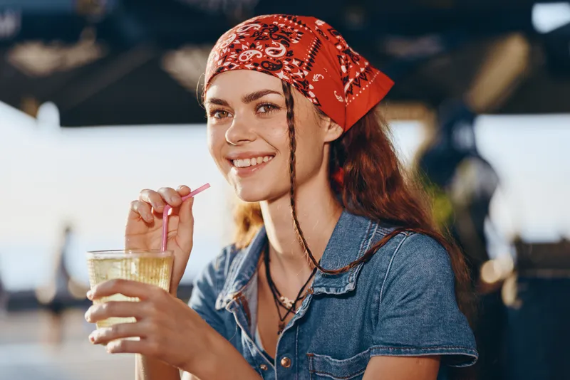 lifestyle woman in modern boho-western denim street style with red bandana smiling and holding a drink, film color tone, relaxed outdoor atmosphere.