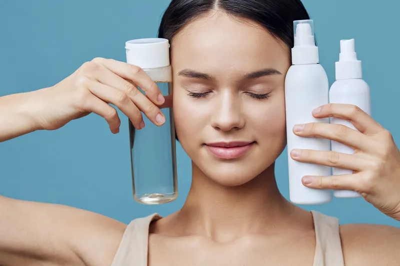 Young woman holding skincare products with a serene expression against a blue background, promoting healthy skin and beauty routines