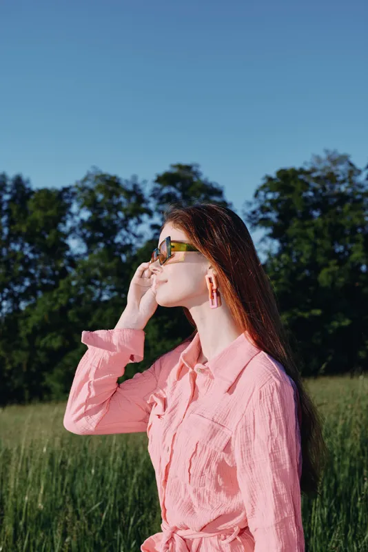 Young woman in pink shirt and sunglasses enjoying a sunny day in a green field, conveying joy and confidence under a clear blue sky