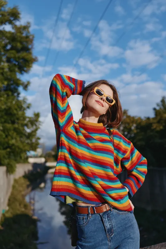 Woman in her 20s wearing a rainbow sweater and sunglasses, casual outfit with jeans, smiling and hands behind head in outdoor urban nature setting for relaxed lifestyle and fashion portrait.
