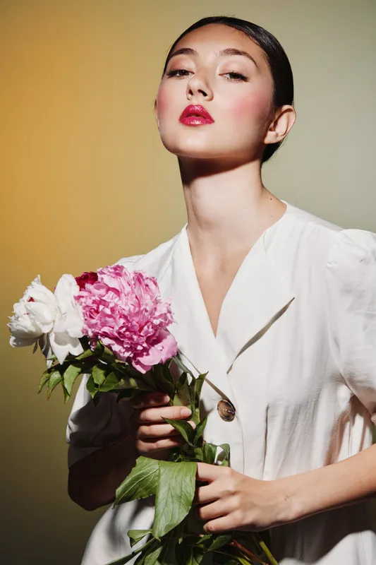 Elegant woman in a white dress poses holding pink and white flowers. Dramatic studio lighting highlights her glossy red lips and smooth skin. Concept for beauty, fashion, creativity.