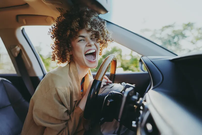 Joyful woman driving a car, laughing with open mouth, warm retro color tones, candid lifestyle moment, playful energy, casual fashion, sunny mood and happiness.