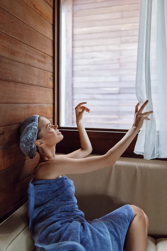 Relaxed woman wrapped in blue towel and shower cap sitting indoors by wooden wall and window, reaching gently for white curtain with soft natural light.