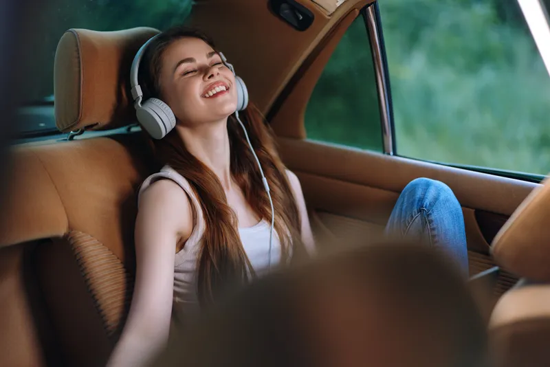 Young woman enjoying music in a car, smiling and relaxed, wearing casual clothes and headphones, green nature background, summer vibe.