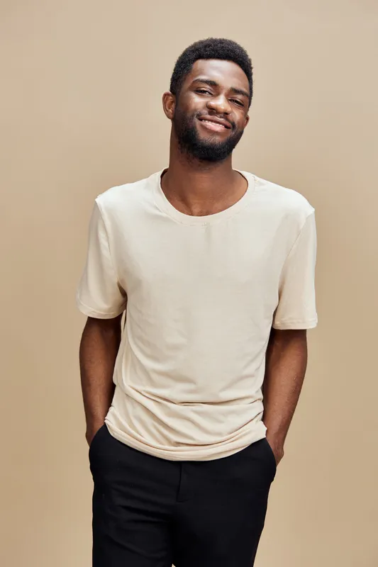Smiling African American man in a casual beige t shirt and black pants against a neutral background, exuding confidence and approachability