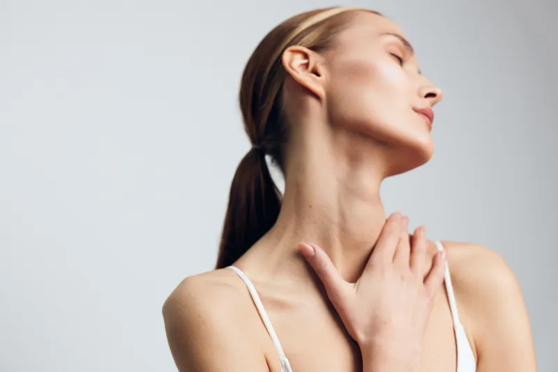 Young woman with blonde hair tied back gently touches her neck and looks upward with closed eyes, expressing calmness and serenity in a minimalistic setting.