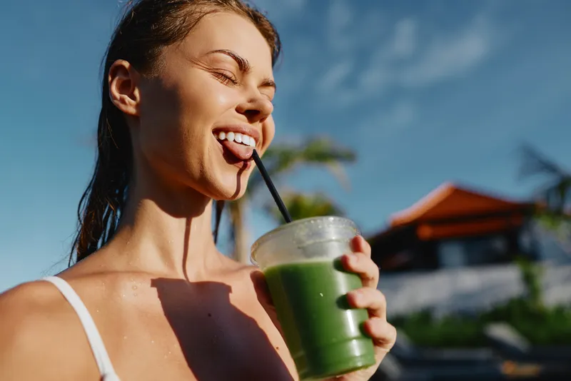Bright woman outdoors, sipping a green smoothie through a straw, sunlit expression, casual tank top, refreshing moment, palm trees and blue sky in the background, relaxed lifestyle vibe
