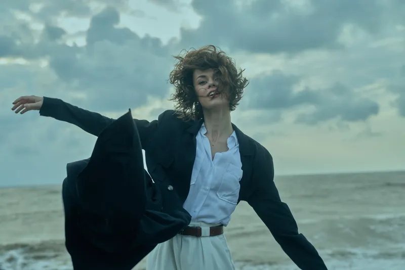 Windy beach fashion moment featuring a woman in a blazer and white shirt, coat sweeping in the breeze, sea horizon behind for a bold outdoor lifestyle and dynamic movement and energy