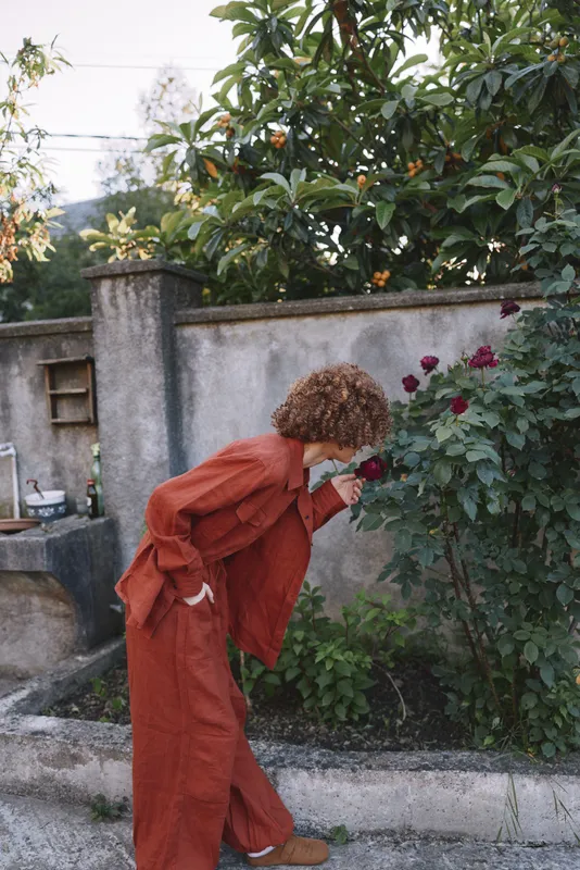 Curly haired woman in a red orange outfit leans toward a flowering rose bush in a garden, capturing a candid outdoor lifestyle moment with natural light and gentle curiosity
