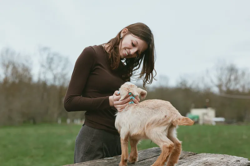 Woman smiles as she pets a goat kid on a rock in a grassy field, a candid lifestyle moment, gentle interaction, natural light, rustic setting, joyful connection outdoors