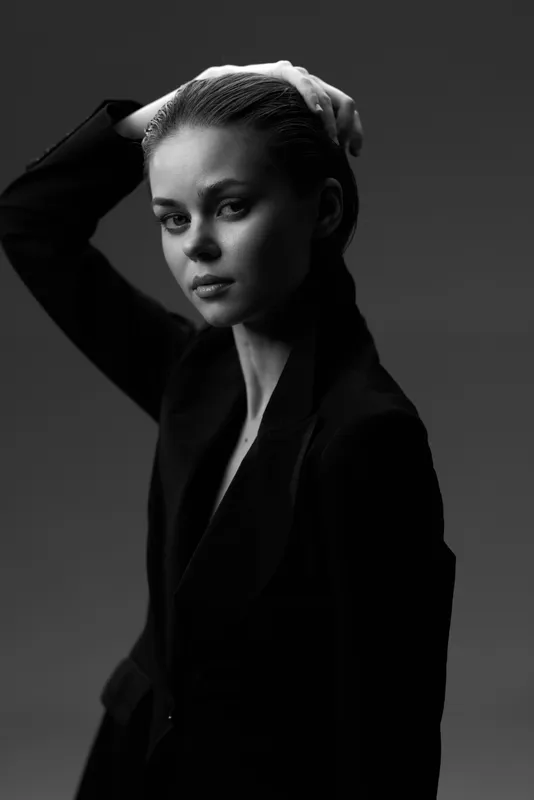 Black and white portrait of a serious woman in a dark coat with braided hair, posing with a hand on her head in a studio setting.