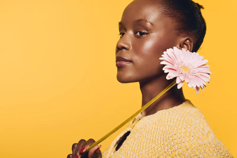 Portrait of a young black woman with dark skin holding a pink flower on her shoulder. She wears a yellow sweater against a yellow background. Concept of beauty, nature, and self-confidence.