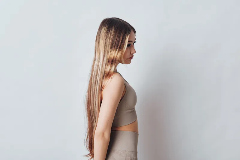 Portrait of woman with long hair standing sideways against plain background wearing casual sleeveless top and pants, natural light and calm mood in studio setting.