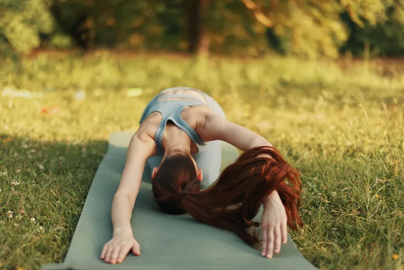 Young woman practicing yoga outdoors on a green mat, embodying tranquility and balance in a sunny park setting with natural greenery