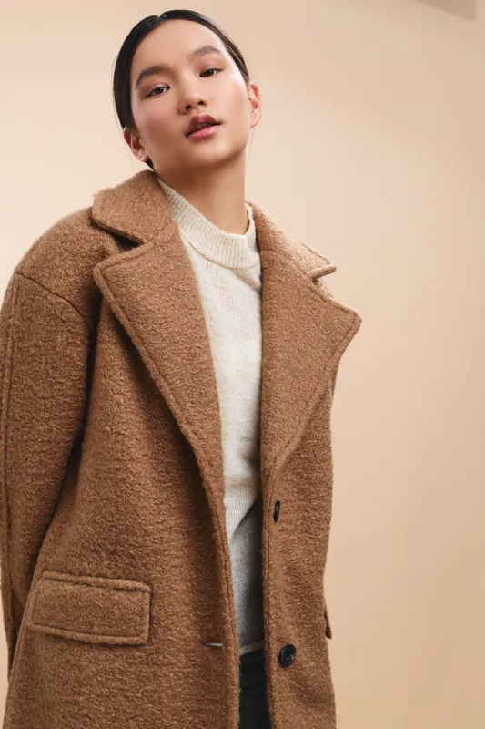 Asian woman wearing a warm coat stands in a neutral studio, presenting a poised, stylish look with soft lighting, conveying elegance and contemporary fashion.