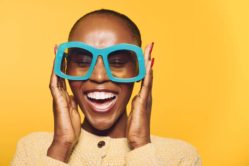 Happy African woman with dark skin wearing large blue sunglasses and yellow sweater smiles with closed eyes on bright yellow background. Concept of joy, fashion, and summer style.
