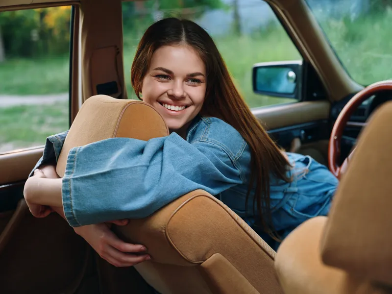 A cheerful young woman in a denim jacket leans over the car seat, smiling warmly, reflecting a joyful summer vibe in a natural outdoor setting.